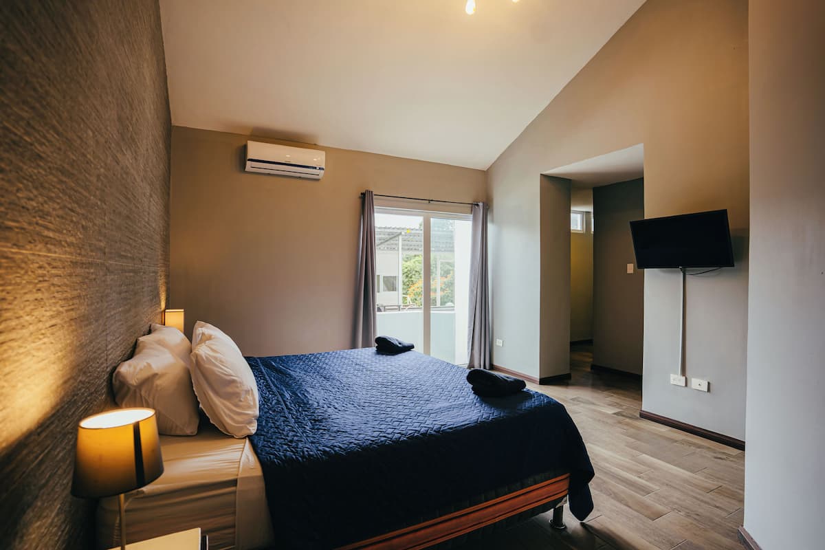 Een slaapkamer met een bed, een bedlamp, een airconditioner, een aan de muur bevestigde televisie en uitzicht op het omliggende landschap door een raam.