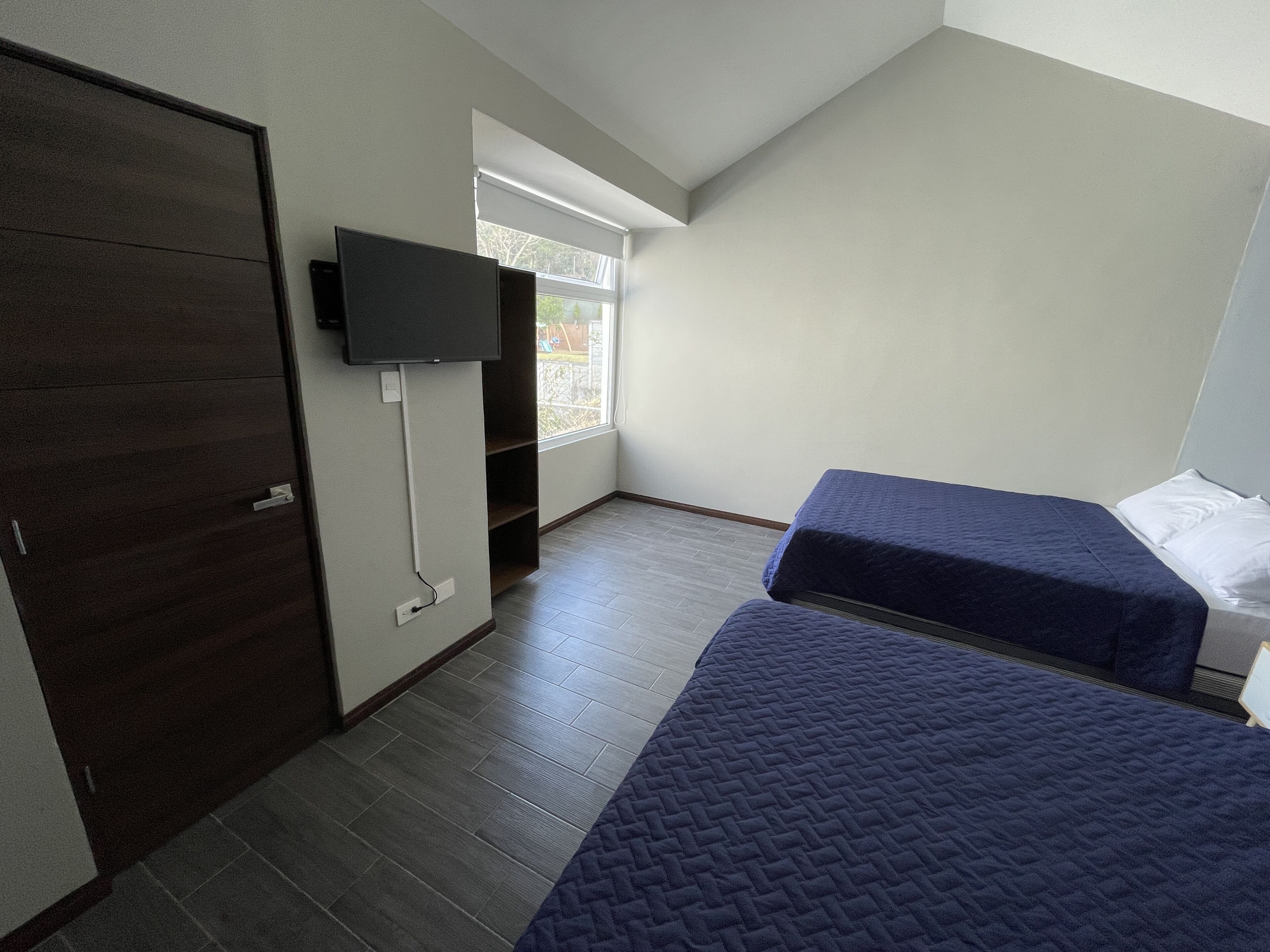 A small, modern room with two single beds, a TV mounted on the wall, and a window providing natural light.