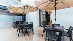 Een moderne eetruimte met houten tafels en stoelen, grote parasols en een glazen wand met uitzicht op een stadsgezicht.