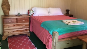 Una cama de madera con colcha roja y verde, una mesita de noche de madera con cajones, una cesta de mimbre y un espejo en la pared.