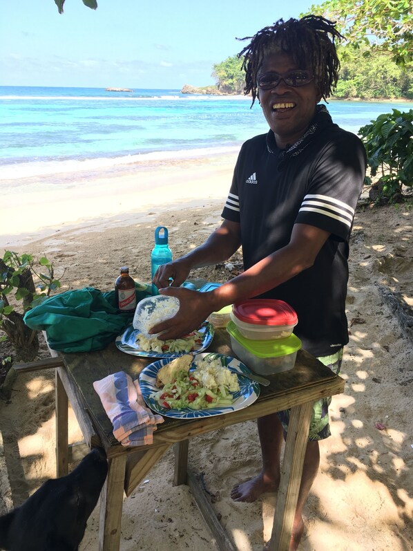 Une personne prépare des aliments sur une table près d’une plage, avec vue sur l’océan.