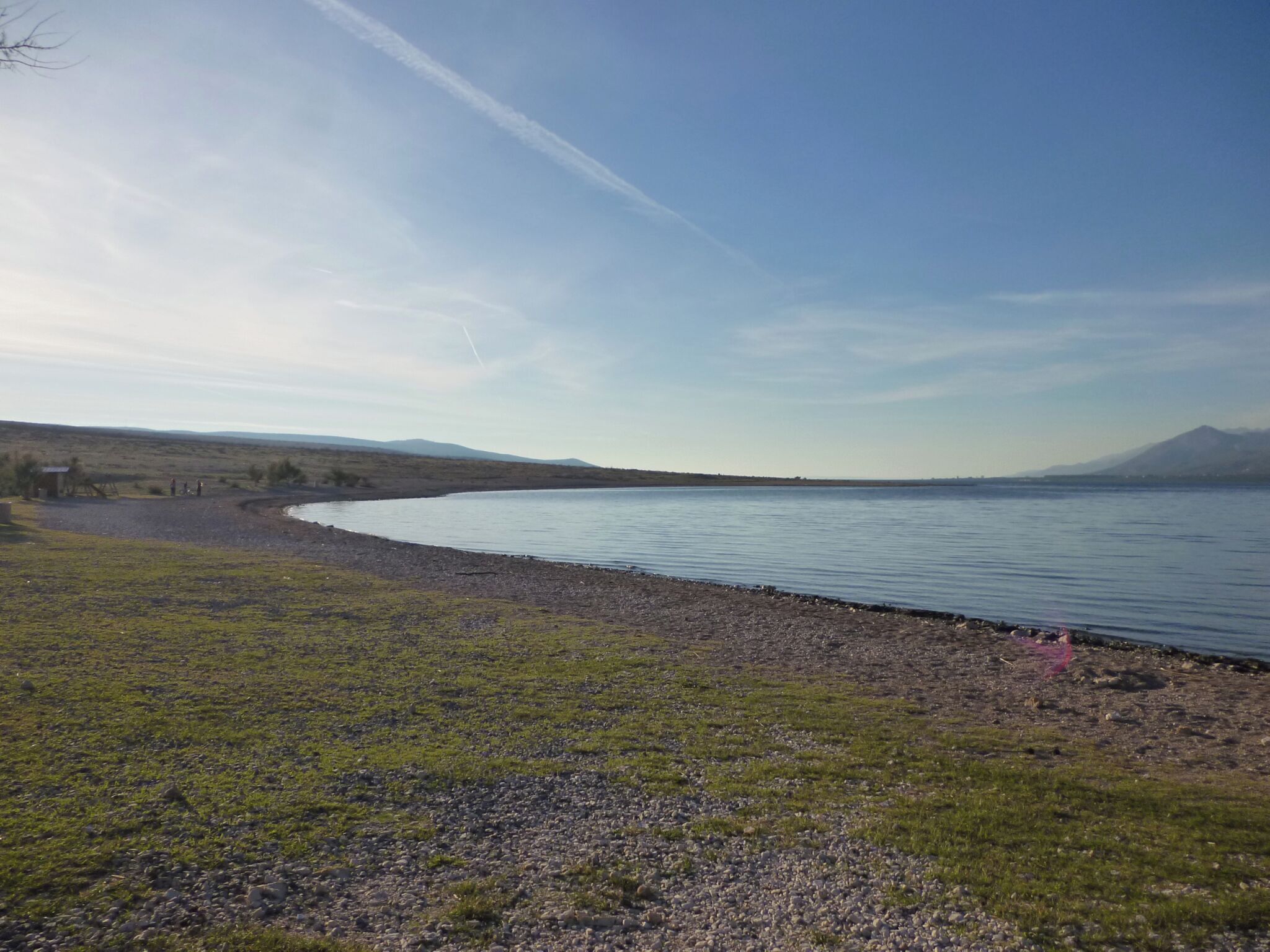 A lakeside view with a grassy shore, a small building, and distant mountains.