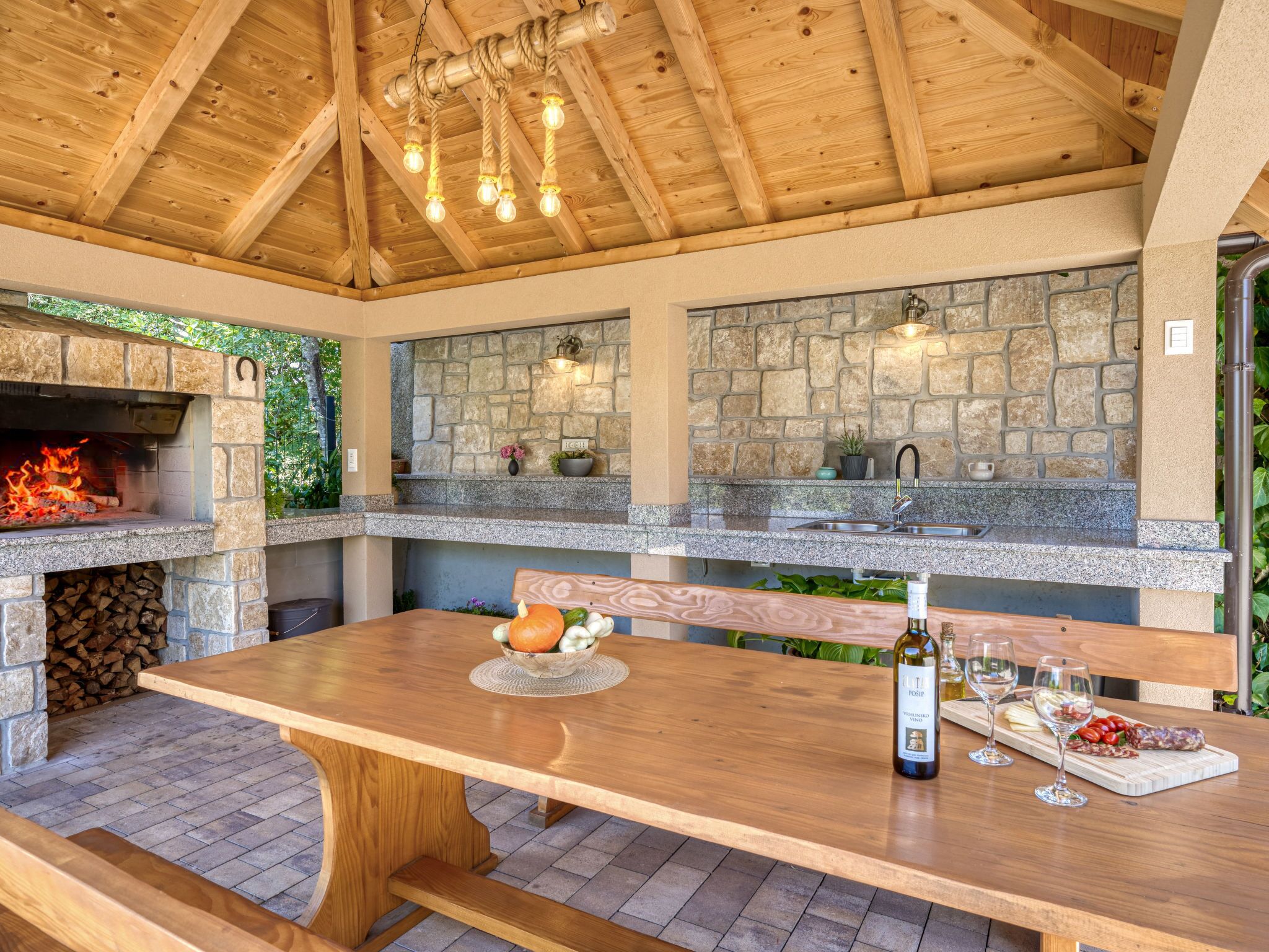 An outdoor kitchen with a stone backsplash, a built-in grill, and a wooden table with a bottle of wine and glasses.