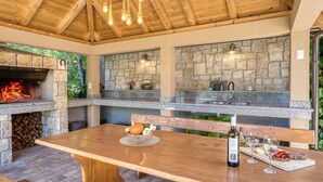 An outdoor kitchen with a stone backsplash, a built-in grill, and a wooden table with a bottle of wine and glasses. - Vacation home Matea by Interhome (Krk/Dobrinj)