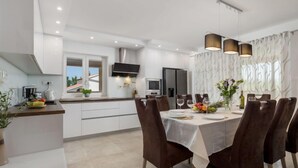A modern kitchen with white cabinets, a dining area with a table set for four, and a large window with curtains. - Vacation home Matea by Interhome (Krk/Dobrinj)