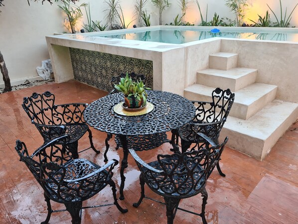 A round metal table with four chairs, a potted plant, and a small pool with steps leading to it.