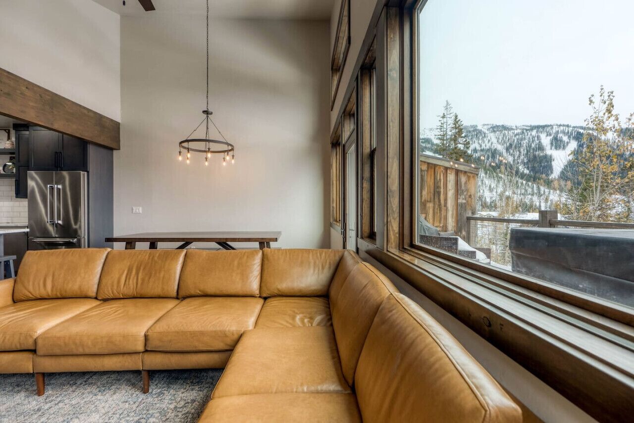 Una sala de estar moderna con un amplio sofá de cuero, una mesa de comedor y vistas a un paisaje montañoso nevado a través de grandes ventanales.