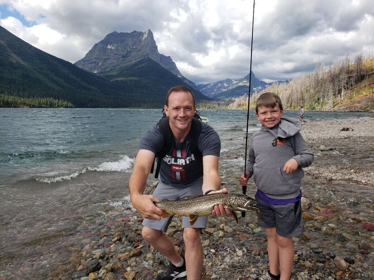 Two people holding a fish by a river with mountains in the background.