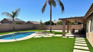 Ein Garten mit Schwimmbad, Kunstgrasanlage, einer Terrassenfläche mit Gartenmöbeln und einer überdachten Sitzecke.