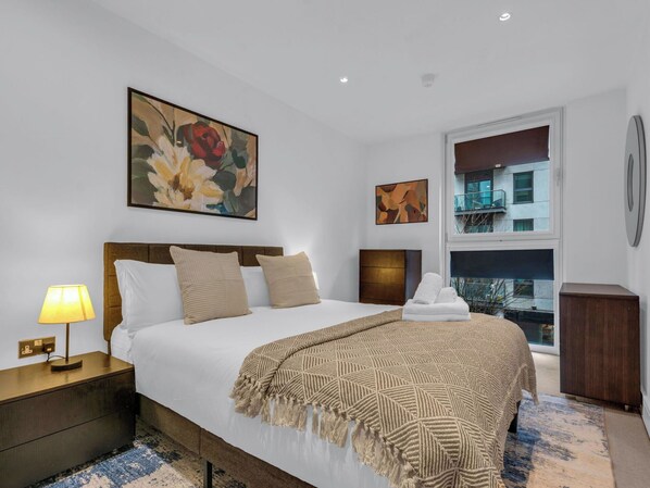 A hotel room with a bed, a nightstand, a lamp, a window with a view, and artwork on the walls.