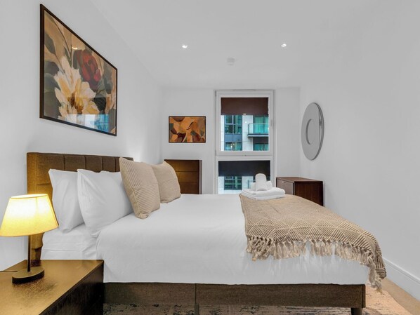 A hotel room with a bed, a nightstand, a lamp, a window with a view of buildings, and artwork on the walls.