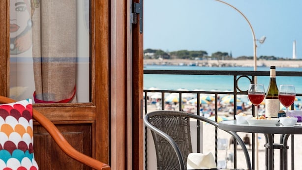 Un balcon avec vue sur la plage, une table avec des verres à vin et une bouteille, et une chaise agrémentée d’un coussin coloré.