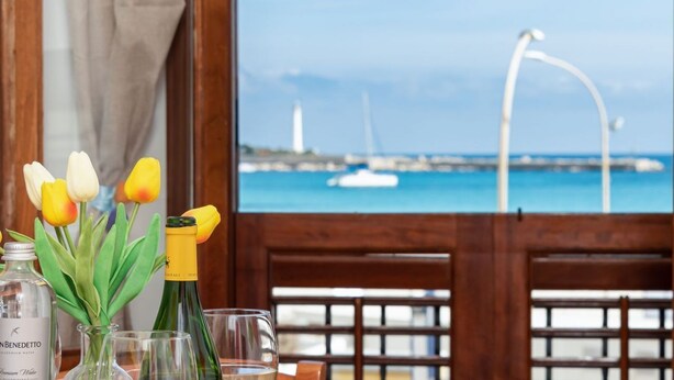 A table with a bottle of water, a glass, and a vase of tulips by a window overlooking a harbor with boats and a bridge. - Beach Apartments - Two-Room (San Vito Lo Capo)