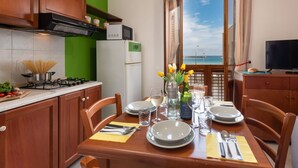 A dining area with a table set for two, a kitchen with a stove and cabinets, and a view of the sea through a window. - Beach Apartments - Two-Room (San Vito Lo Capo)