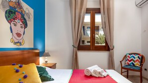 A hotel room with a bed, a chair, a window with curtains, a painting on the wall, and a view of the outside. - Beach Apartments - Two-Room (San Vito Lo Capo)