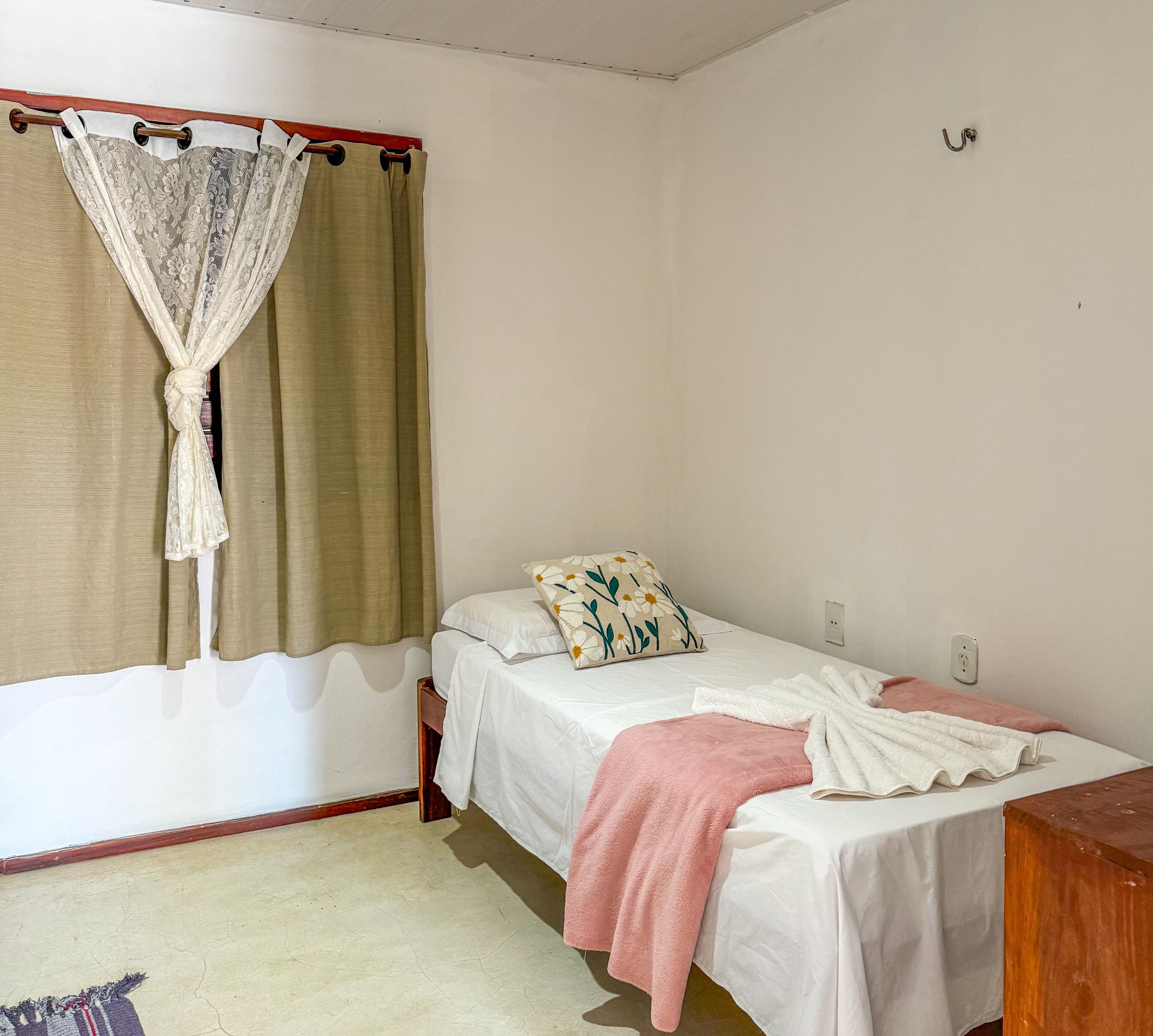 Une chambre avec un lit simple, une tête de lit en bois, un rideau beige et un mur blanc.