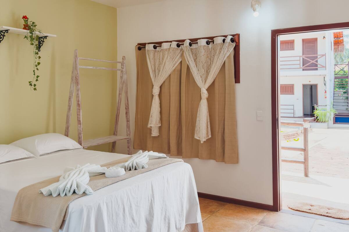 A hotel room with a bed, a wooden ladder, a window with curtains, and a view of the outside.