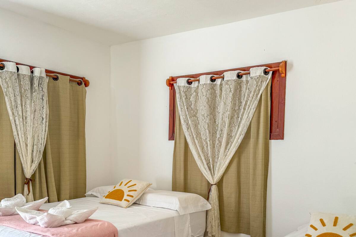 A hotel room with two beds, each with a white bedspread, a pink blanket, and towels. There are curtains with a floral pattern and a wooden curtain rod.