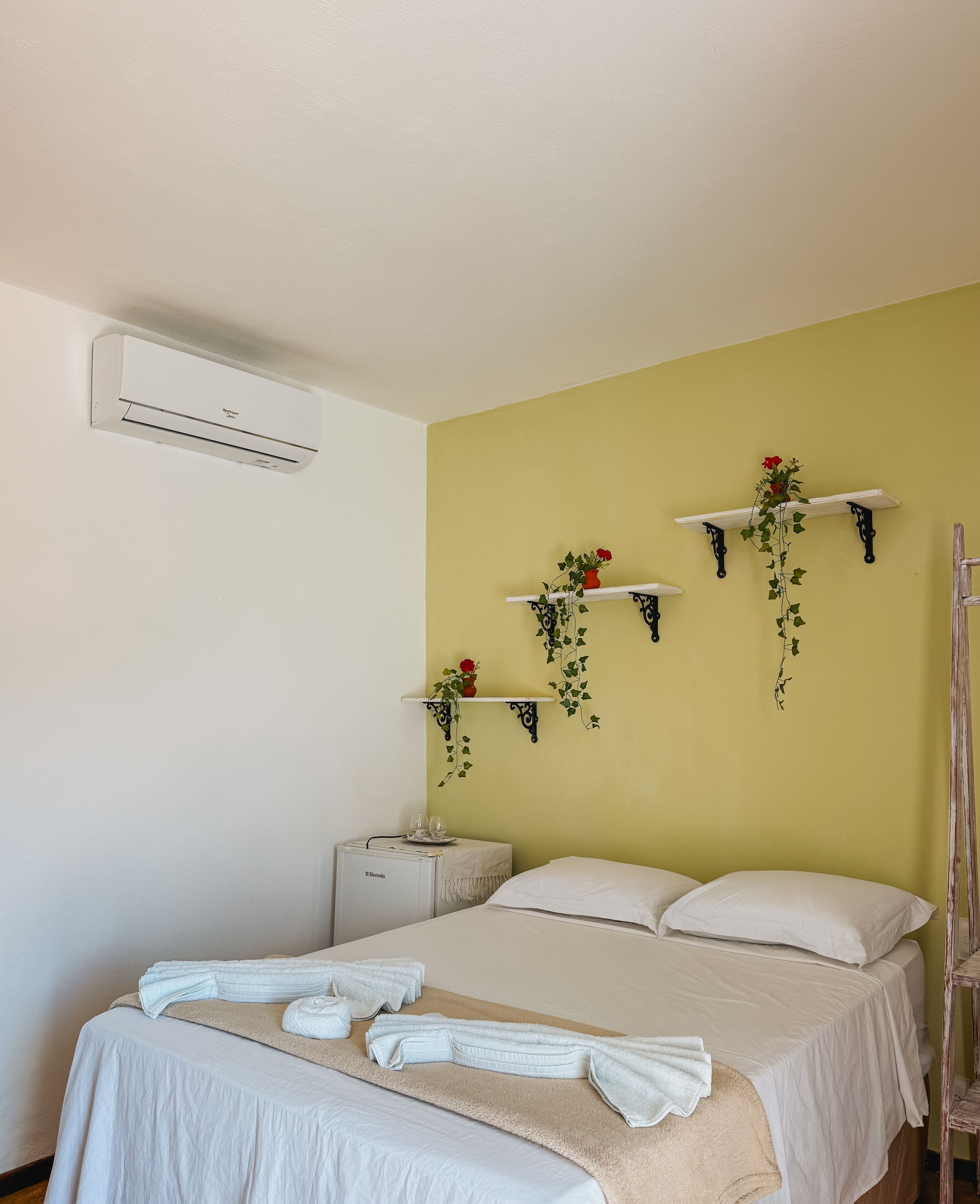 A hotel room with a bed, white towels, a minibar, and wall-mounted shelves with plants.