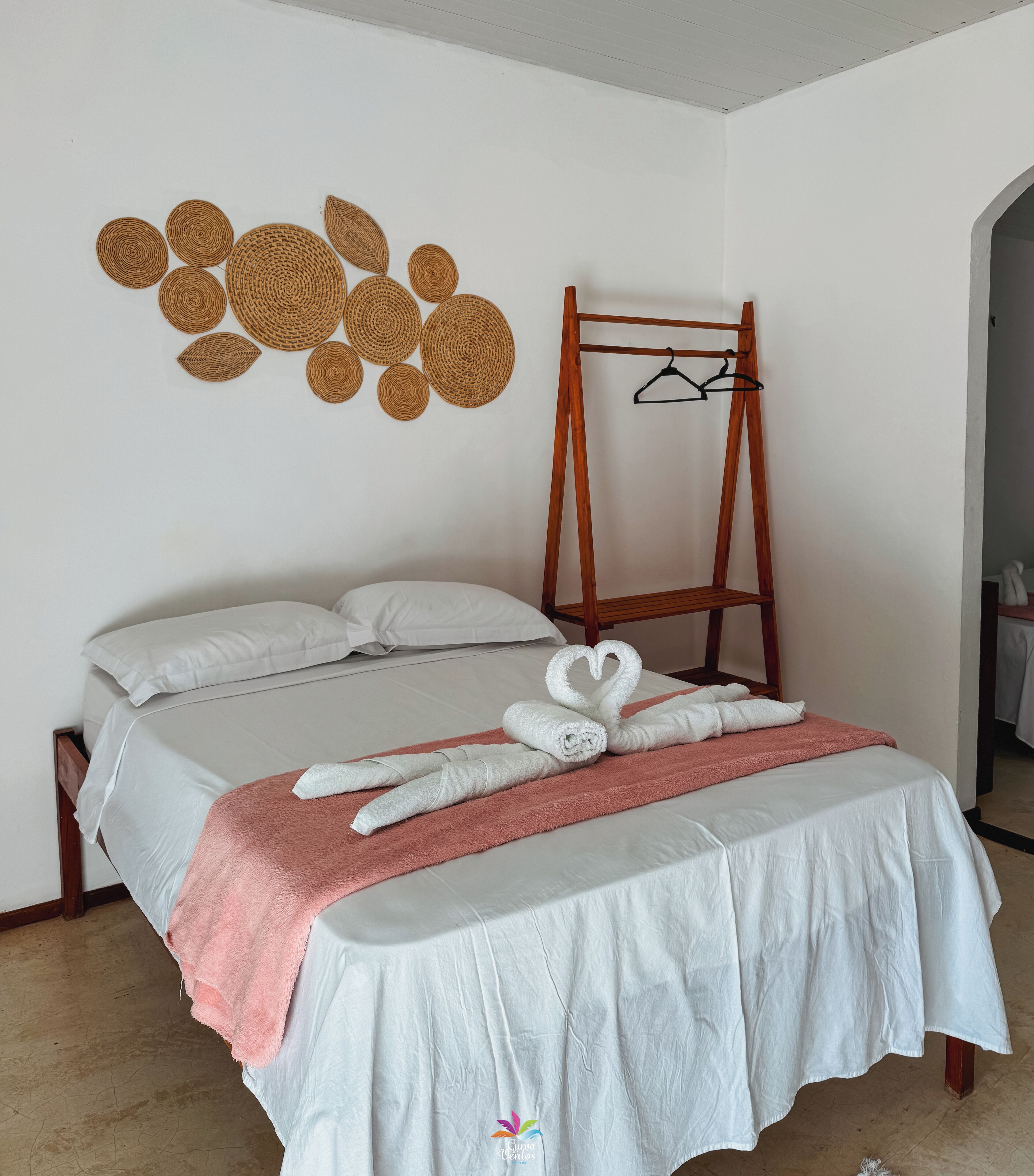 Ein ordentlich bezogenes Bett mit weißen Laken, ein rosa Handtuch und eine Schwanenform aus Handtüchern. An der Wand steht ein hölzerner Kleiderhaken und eine gewebte Wanddekoration.