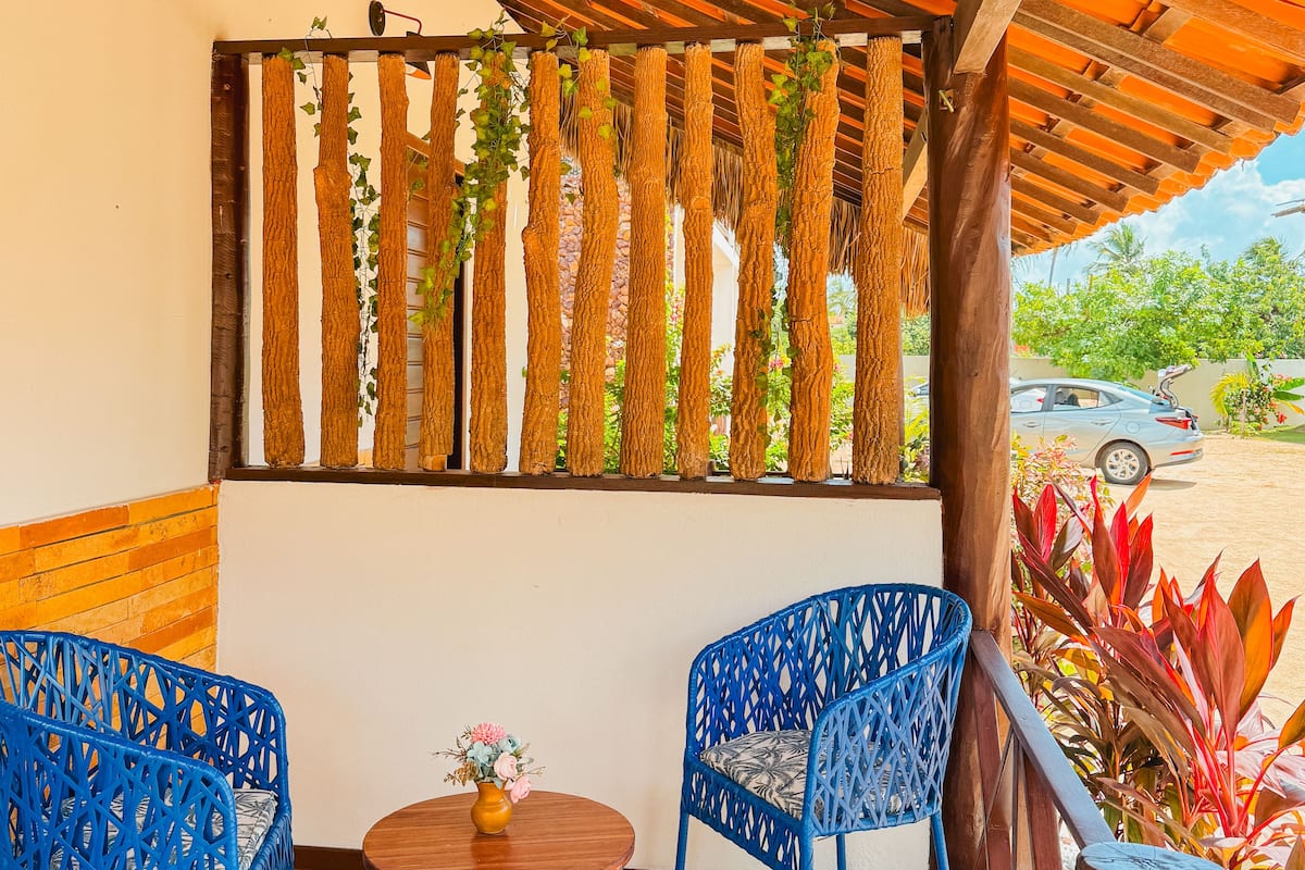 A covered outdoor seating area with blue chairs and a small table, featuring a wooden ceiling and a view of a car and greenery in the background.