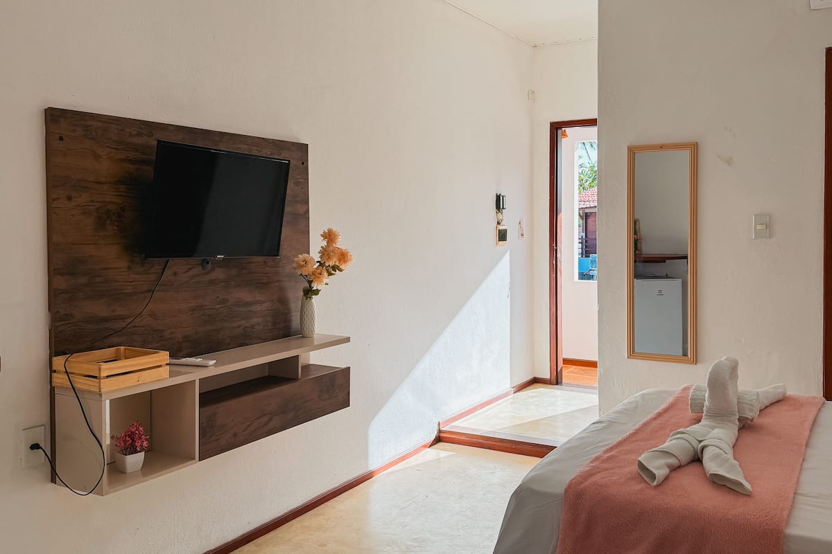 A bedroom with a bed, a TV mounted on a wooden panel, a mirror, and a small shelf with a vase of flowers.