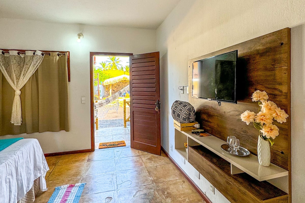 A room with a bed, a TV mounted on a wooden wall, a small table with a vase of flowers, and a door leading to an outdoor area with a hammock and palm trees.