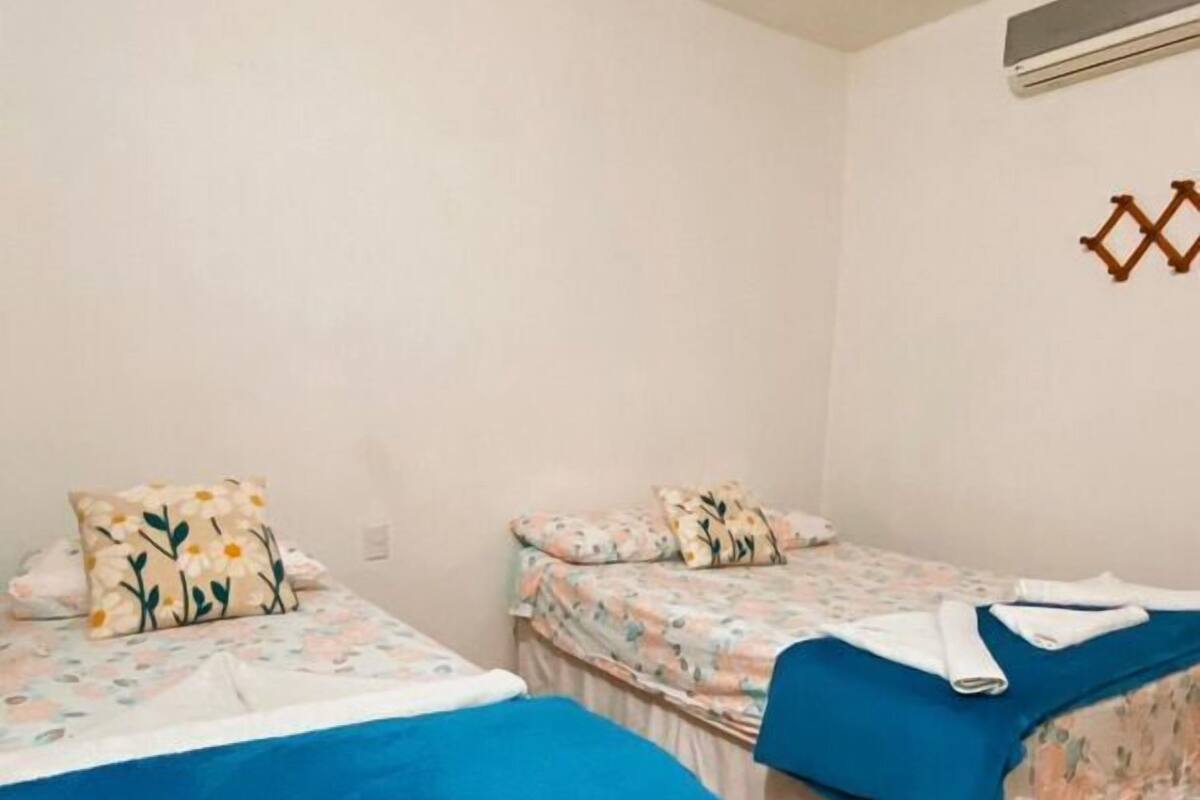 A room with two beds, each with floral bedding and a blue blanket. There is a wall-mounted air conditioning unit and a decorative wall piece.