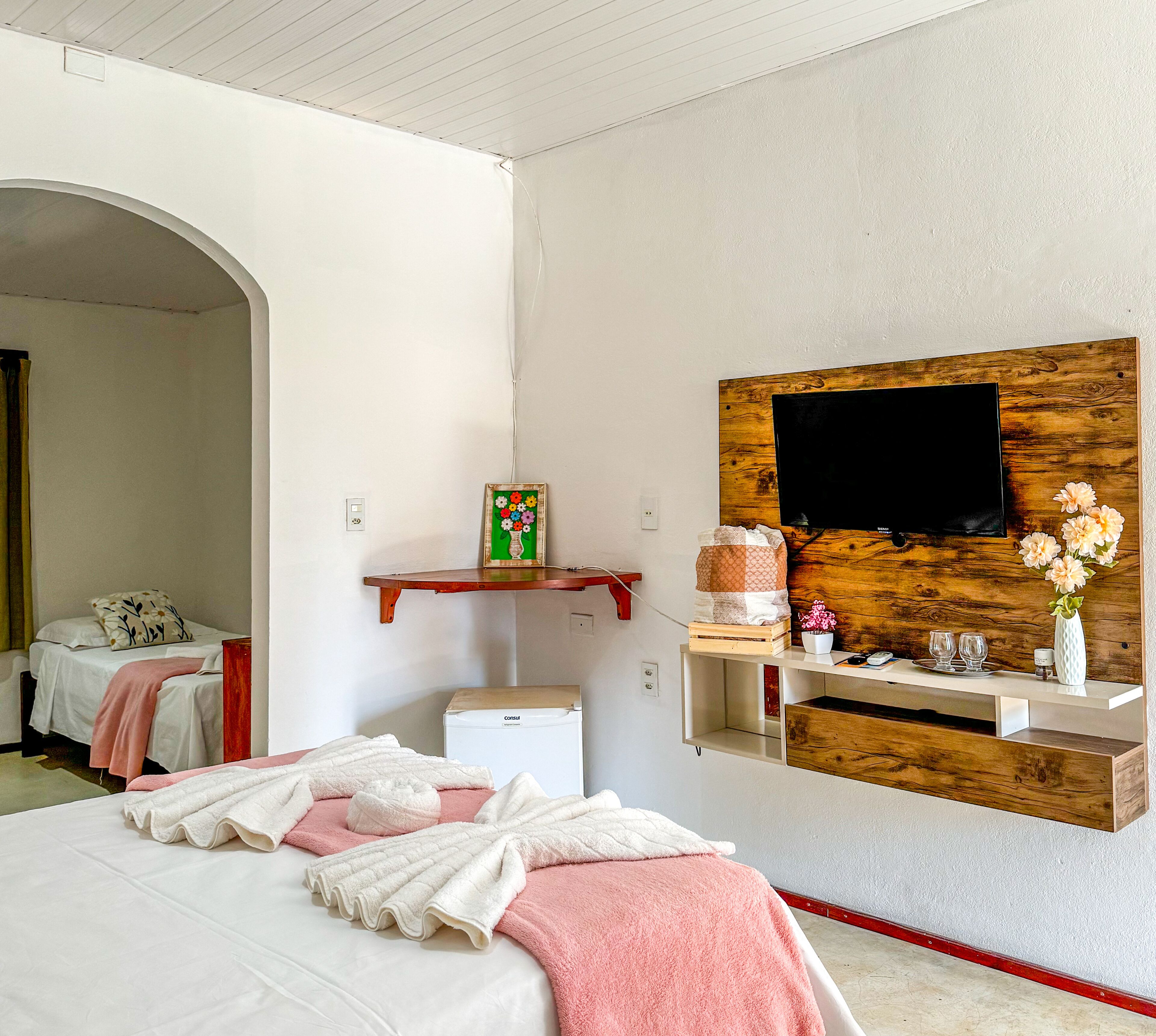 A bedroom with a bed, a TV mounted on a wooden panel, a small shelf with decorative items, and a closet.
