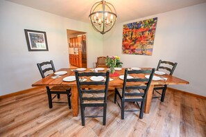 Ein Esszimmer mit Holztisch, gedeckt für vier Personen, umgeben von Stühlen, einer Pendelleuchte und einem farbenfrohen, abstrakten Gemälde an der Wand.