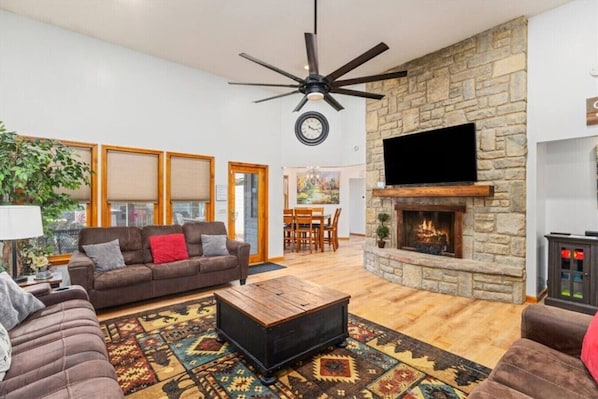 A living room with a stone fireplace, a ceiling fan, a television, and a large window.