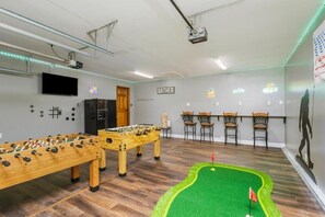 A room with two foosball tables, a ping pong table, a small putting green, and a bar area with chairs.