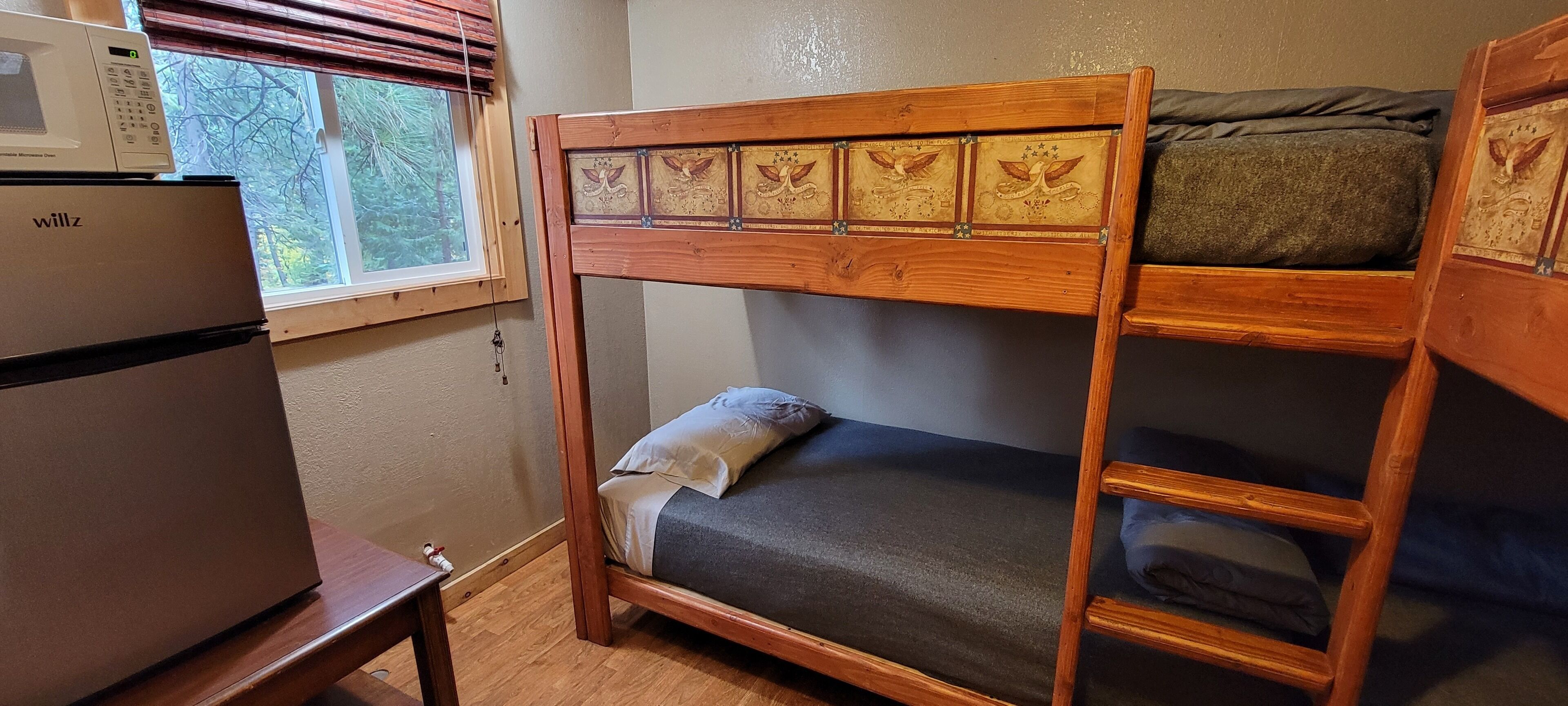 Un lit superposé avec une structure en bois et des panneaux décoratifs, un four à micro-ondes posé sur une petite table et une fenêtre avec des stores.