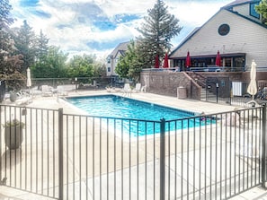 A rectangular pool with a metal fence, surrounded by lounge chairs and umbrellas, with a building in the background.