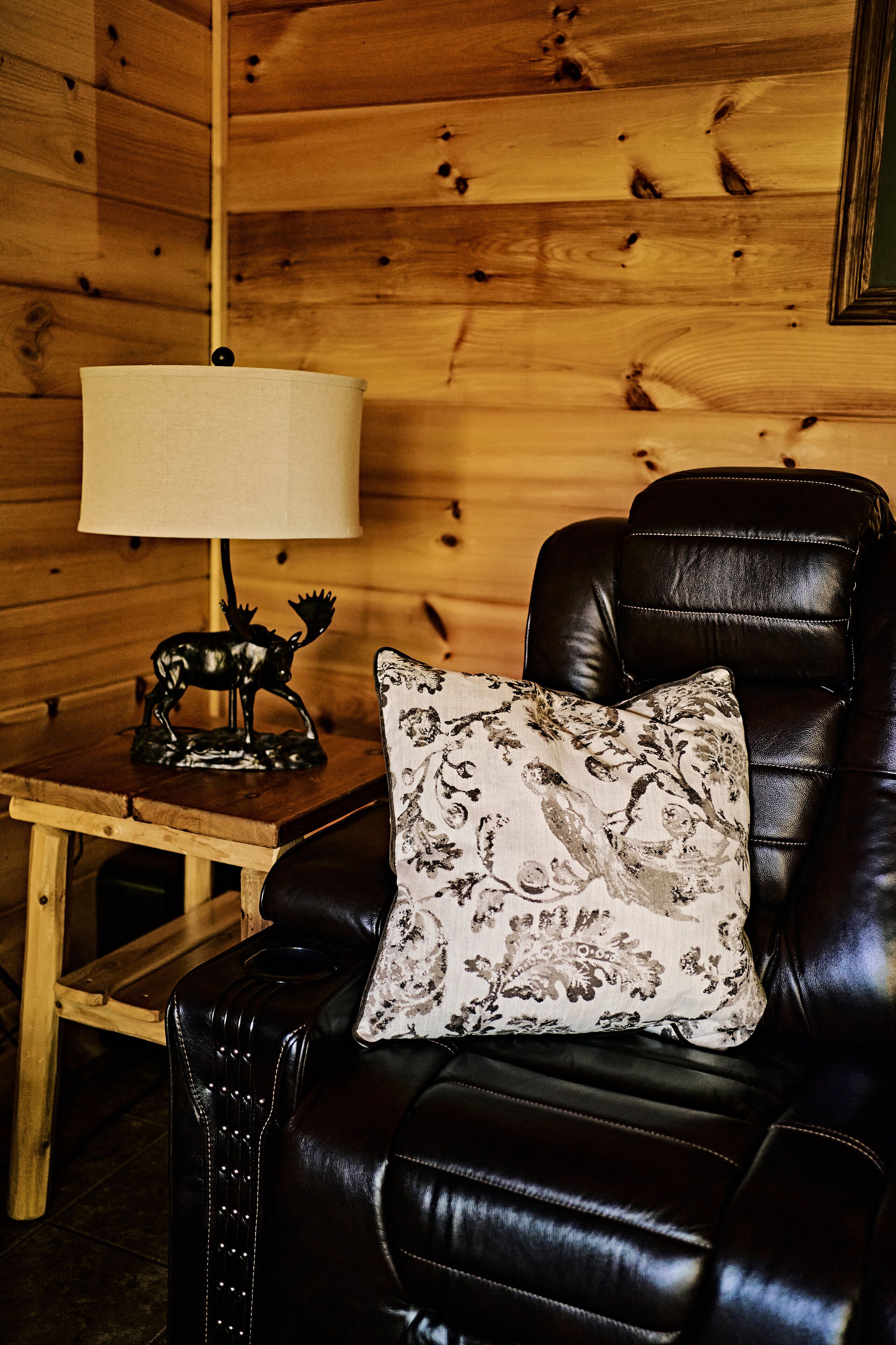 A wooden cabin interior with a leather armchair, a table with a lamp and a moose figurine, and a floral pillow.