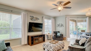 A spacious living room with a large TV, comfortable seating, a ceiling fan, and a view to the outside.