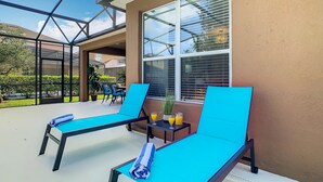 A sunlit outdoor lounge area with two blue lounge chairs, a small table with drinks, and a view of a residential neighborhood.