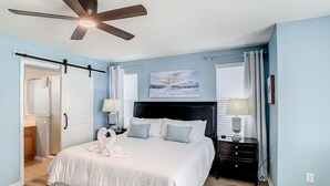 A bedroom with a large bed, a ceiling fan, a nightstand with lamps, and a painting on the wall.