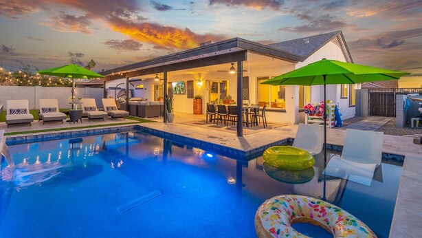 A swimming pool with lounge chairs and umbrellas, a covered patio area, and a sunset view. - Modern Cactus  - Heated Pool and Hot Tub (Peoria)