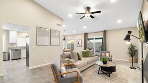 A spacious living area with a sofa, armchair, and coffee table, a ceiling fan, and a kitchen in the background. - Modern Cactus  - Heated Pool and Hot Tub (Peoria)