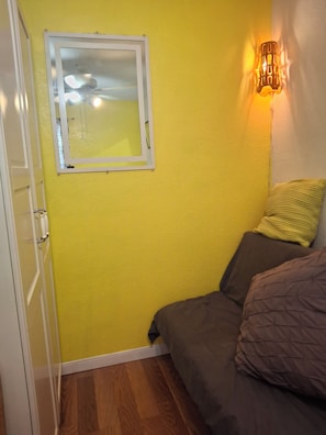 A room with a yellow wall, a window with a white frame, a bed with a brown mattress and pillows, and a wall-mounted light fixture.