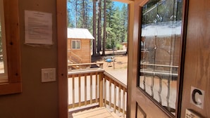 A wooden cabin with a deck, a window with a view of the forest, and a door with a glass panel.