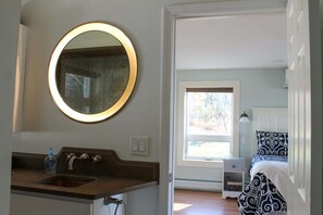 Un baño con un espejo redondo, lavamanos y ventana que da a un dormitorio con cama y mesita de noche.