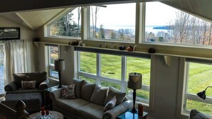 A cozy living room with a large window, a sofa, a chair, a lamp, and a small table with decorative items. - Sunset Waterview Cottage w/Fire Pit Near Acadia (Ellsworth)