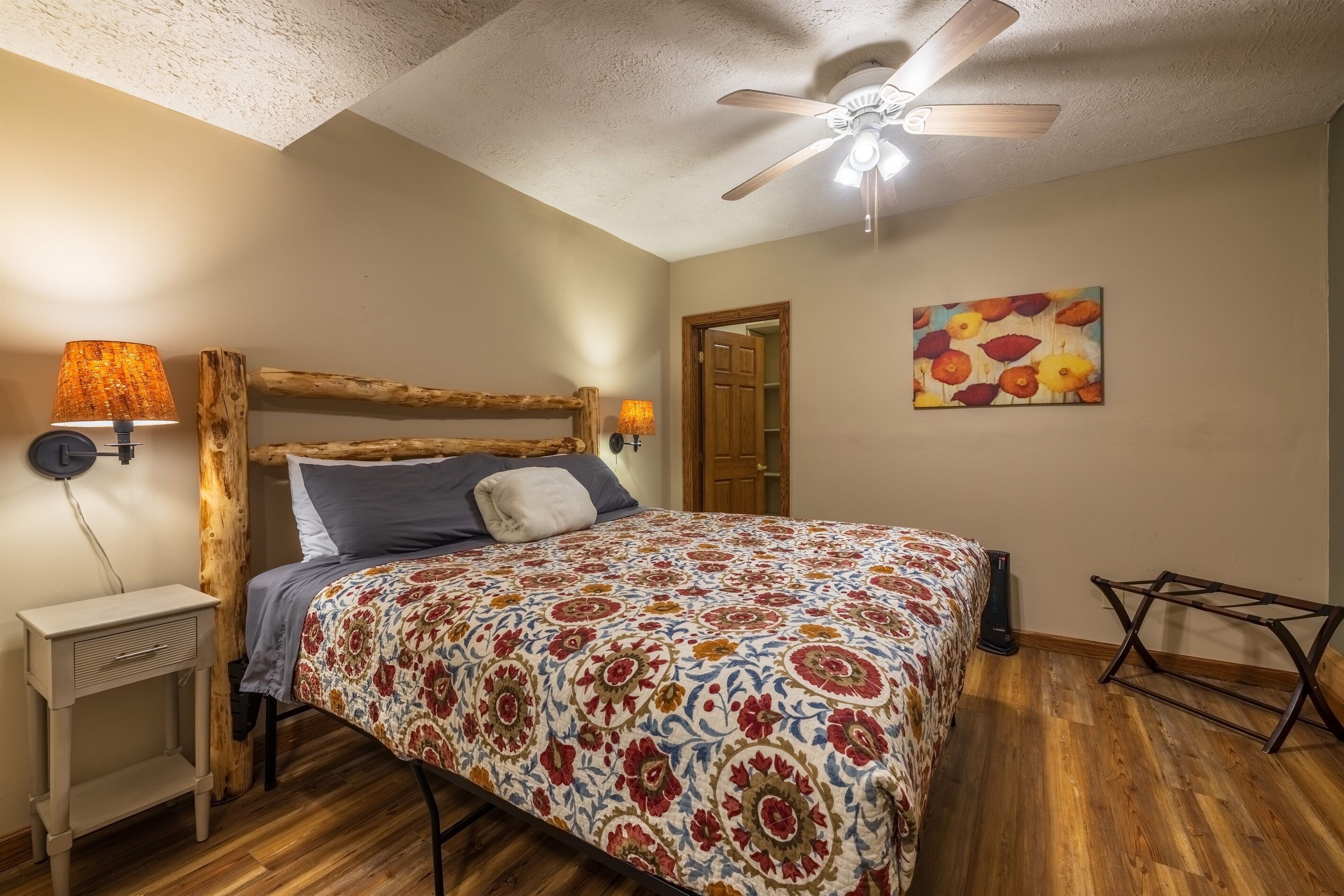 A bedroom with a wooden headboard, a patterned bedspread, a ceiling fan, a nightstand, a wall-mounted lamp, and a door leading to another room.