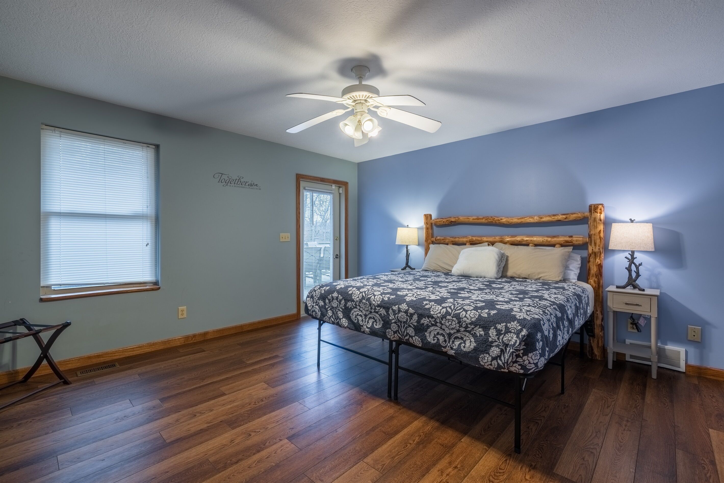 A bedroom with a bed, a ceiling fan, a nightstand, a lamp, a window, and a door.