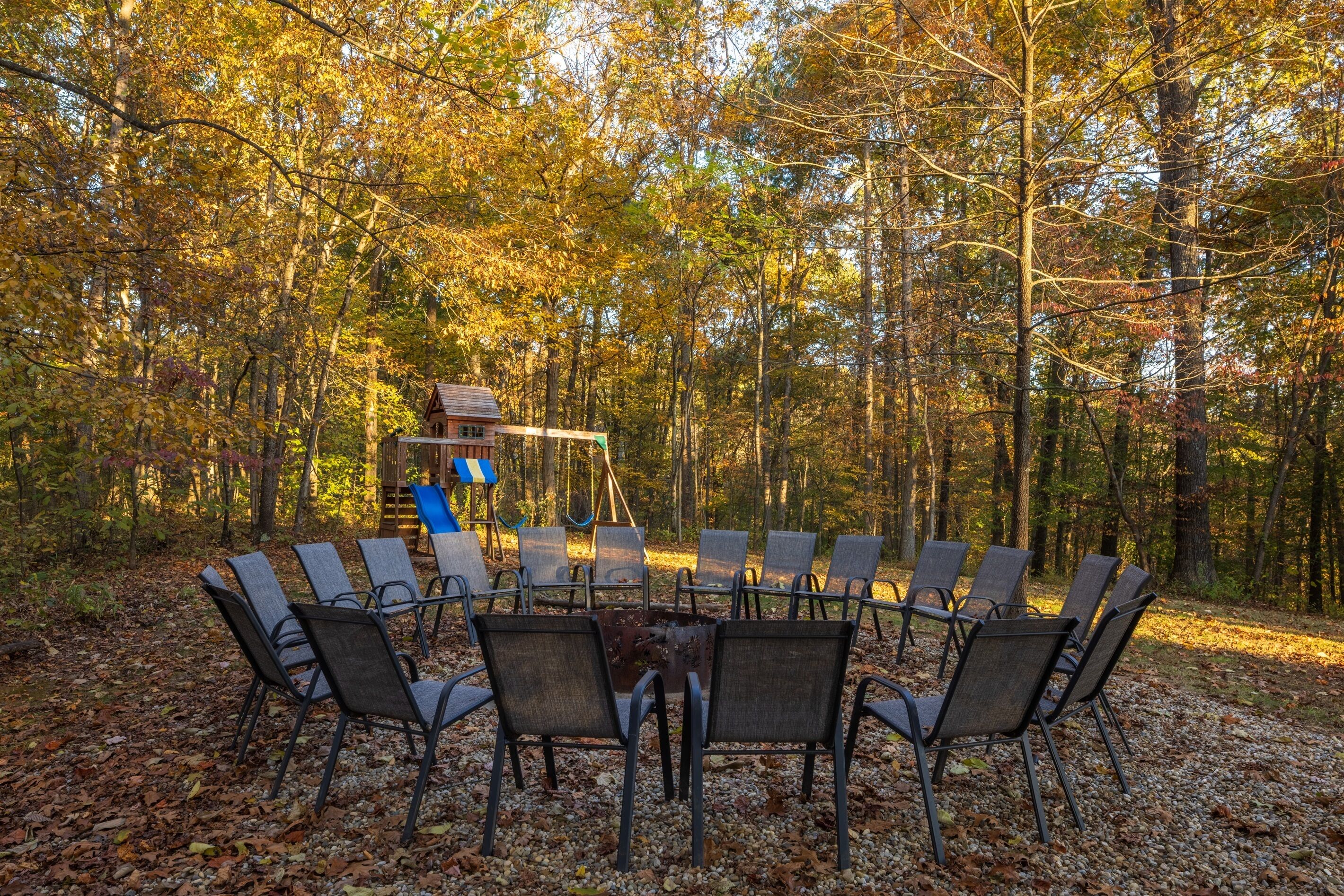 A circular outdoor seating area with chairs and a central fire pit, surrounded by a forest.