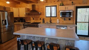 A cabin kitchen with wooden walls, a large island, bar stools, a stove, a refrigerator, and a window with a view of trees.