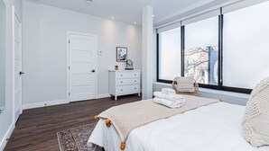 A modern bedroom with a large bed, a dresser, a chair, and a window with a city view. - Vast and perfectly located 3 bedroom flat by Denstays (Montréal)