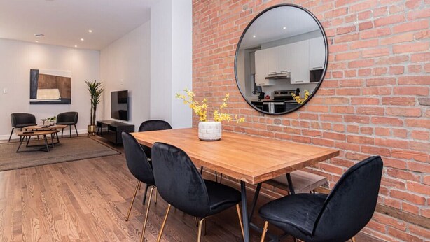 A modern dining area with a wooden table, black chairs, and a round mirror on a brick wall. - Vast and perfectly located 3 bedroom flat by Denstays (Montréal)